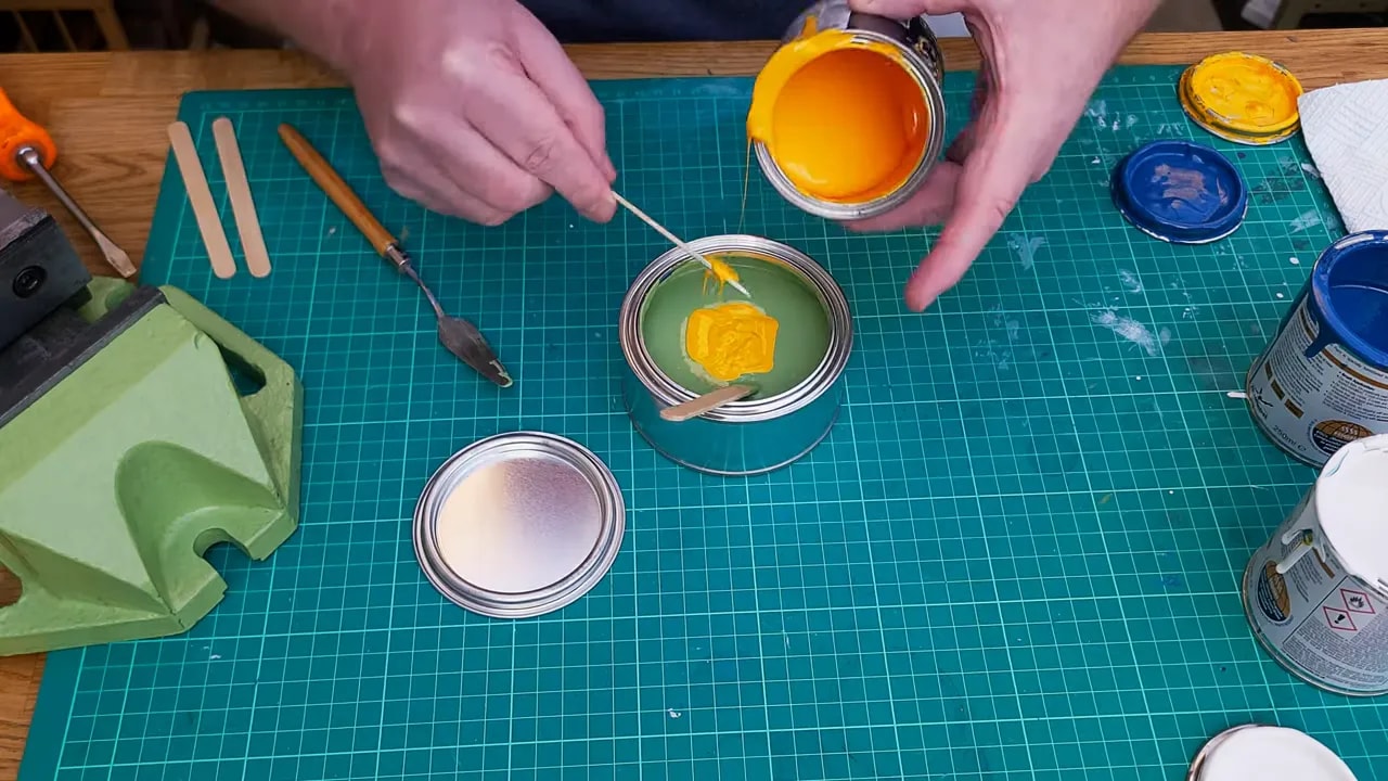 Yellow paint being stirred into pale green Hammerite mixture in a tin on a cutting mat
