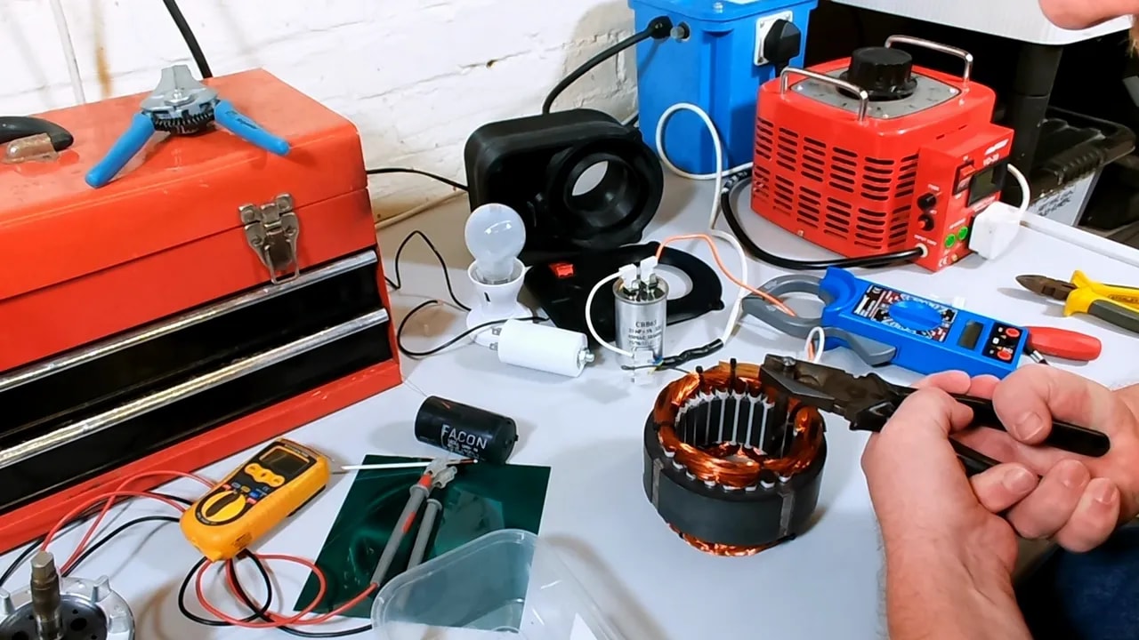Hand holding blue handled pliers with a drill bit near the open stator and red enclosure on the bench