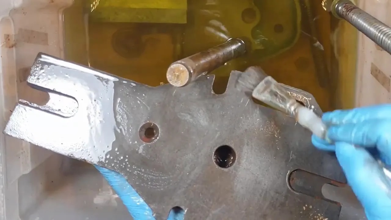 Gloved hands brushing a large grey steel vice plate in yellow-green rust removal solution